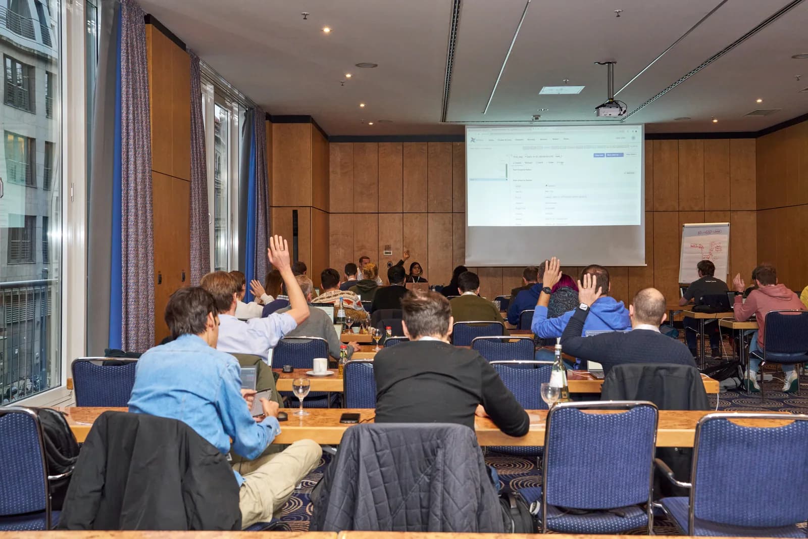A full workshop room of attendees at laptops, several with hands raised, facing a projected Airflow DAG interface.